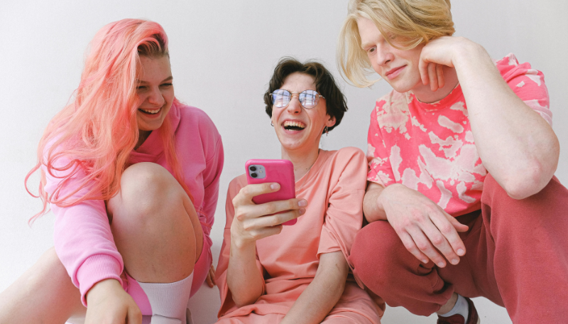 Drei Jugendliche sitzen auf dem Boden und schauen lachend auf den Bildschirm eines Smartphones.