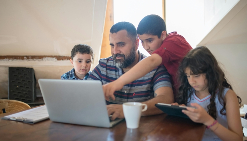 Ein Elternteil mit seinen drei Kindern vor einem Laptop und Tablet