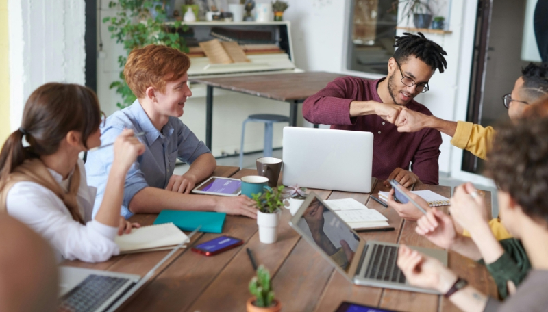 Mehrere Jugendliche sitzen gemeinsam mit einer pädagogischen Fachkraft an einem Tisch. Auf dem Holztisch liegen Laptops, Smartphones und Tablets. Zwei Jugendliche geben sich einen Faustgruß.