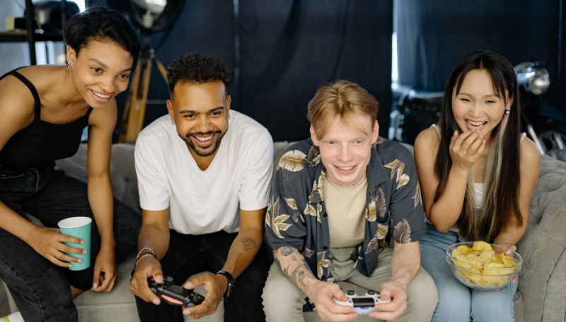 Vier Jugendliche sitzen lachend auf einem Sofa. Zwei von ihnen haben eine Spielekonsole in der Hand.
