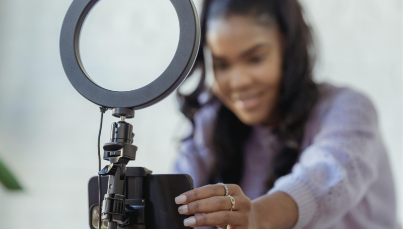 Eine junge Frau steht vor ihrem Smartphone, das an einem Ringlicht befestigt ist und lächelt in die Kamera.